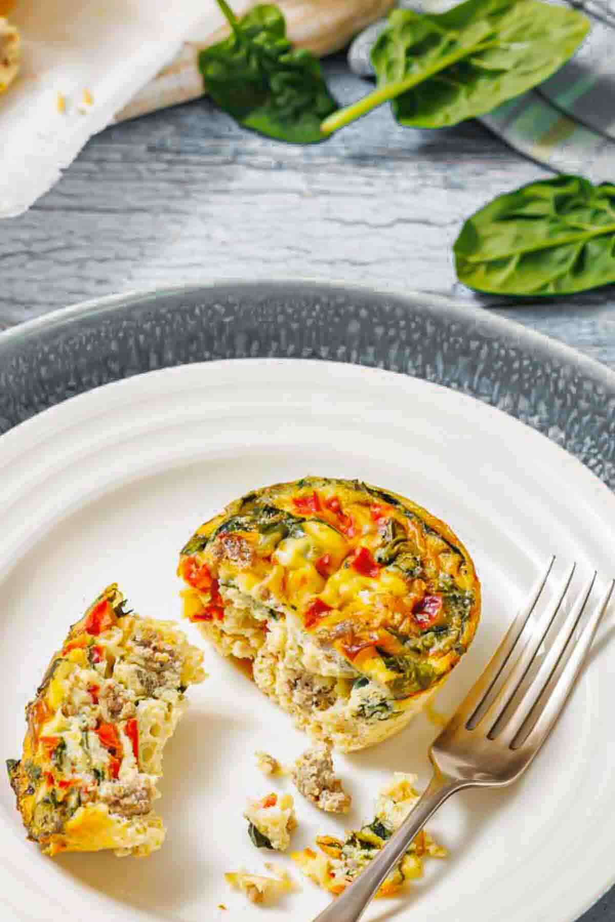 A baked egg muffin with vegetables and sausage sits on a white plate, partially eaten, with a fork beside it and fresh spinach leaves in the background.