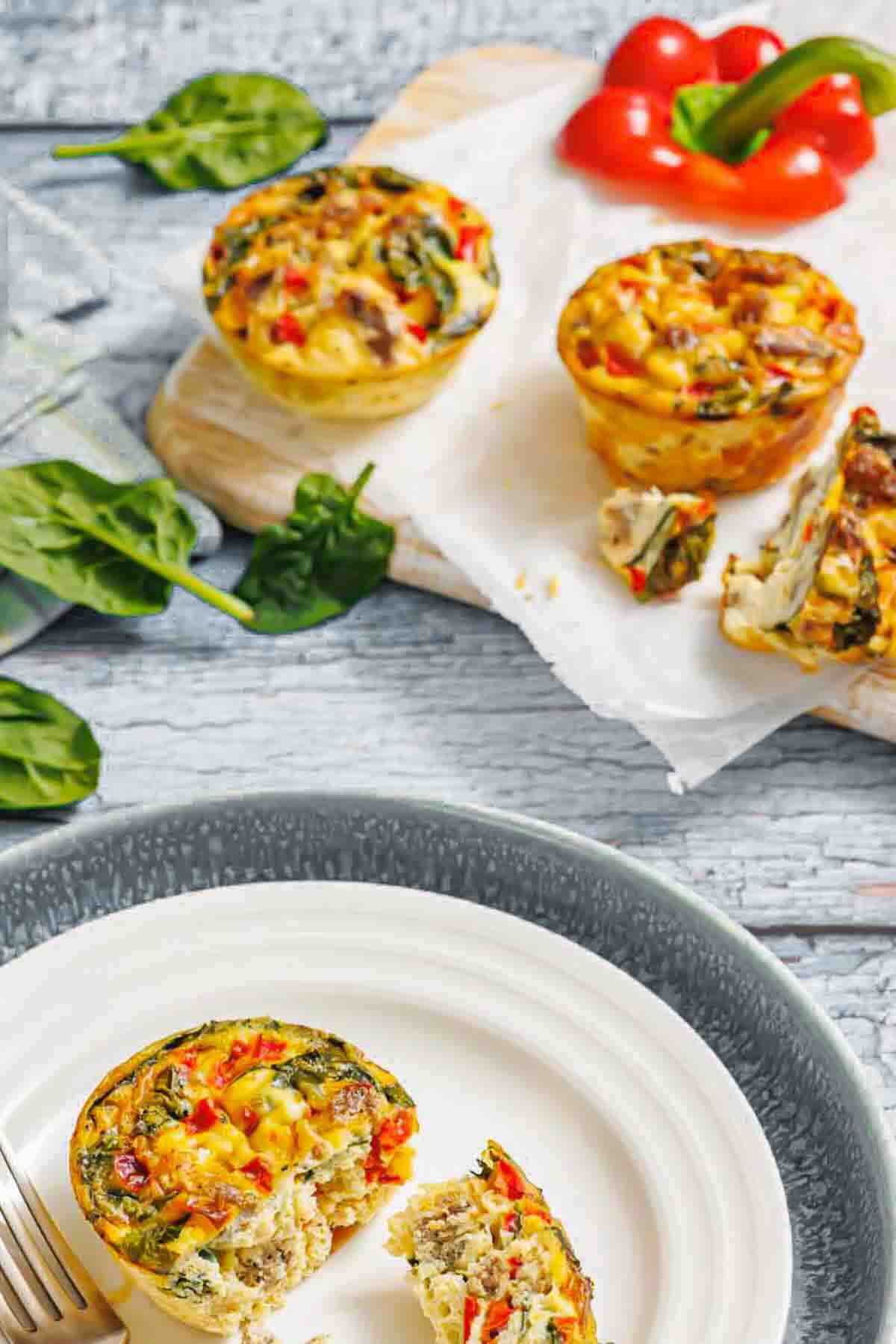 Three mini vegetable frittatas, one sliced open, are displayed on plates and a cutting board with spinach leaves and a red bell pepper nearby.