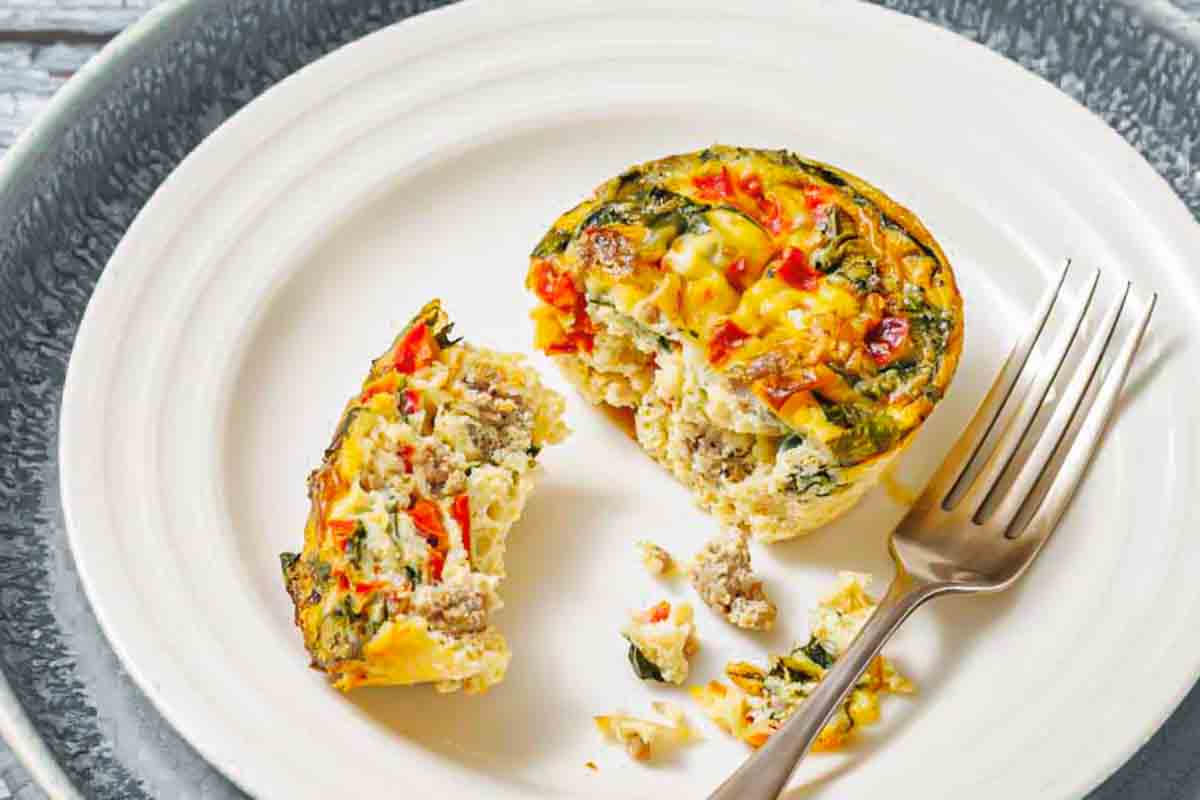 A vegetable and meat egg muffin is cut in half on a white plate with a fork beside it.