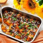 Rectangular dish filled with sweet potato casserole topped with bacon, shredded cheese, and chopped green onions. Spoon rests in the casserole. Sunflowers and orange napkin are nearby.
