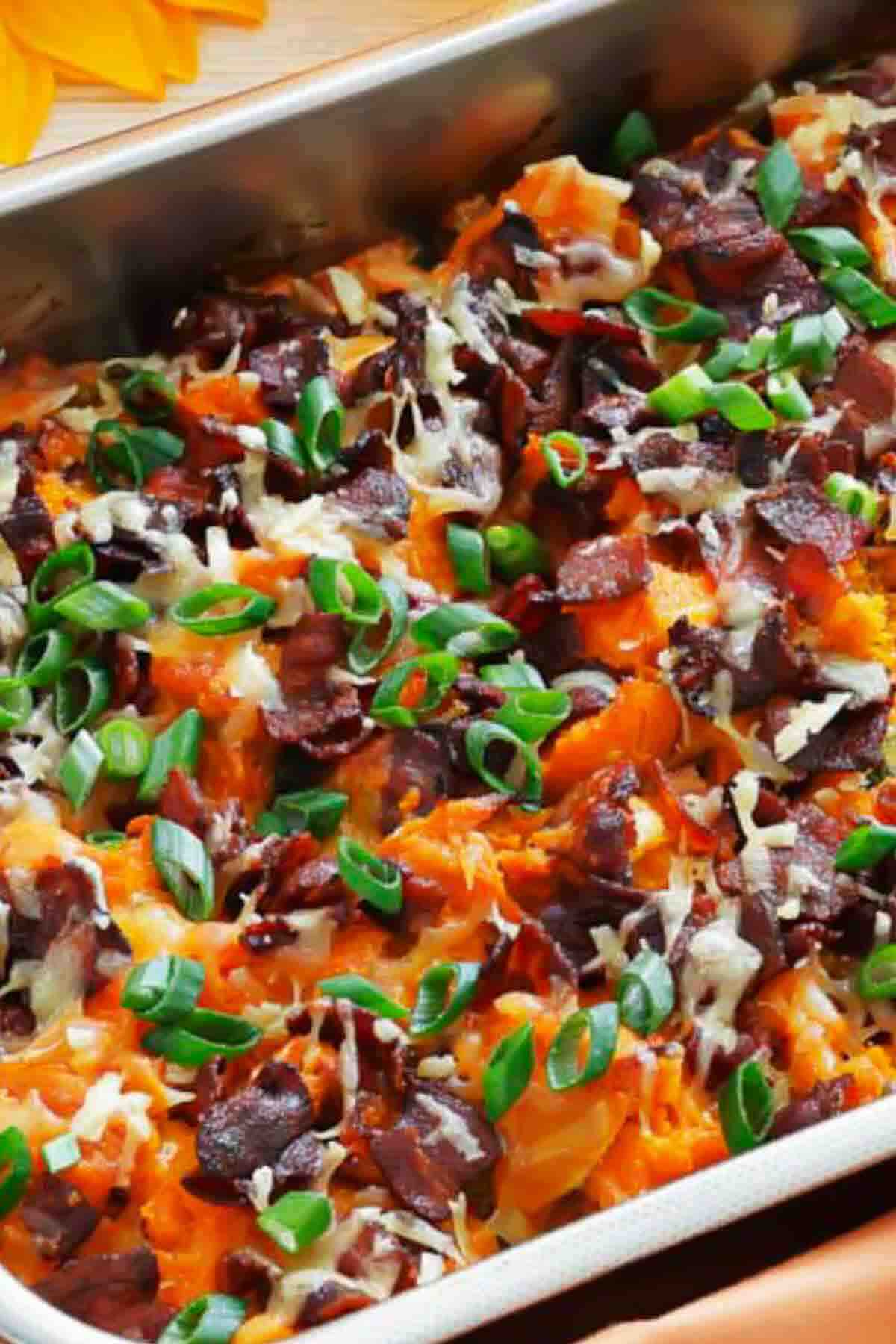A baked casserole topped with crumbled bacon, grated cheese, and chopped green onions in a metal dish.
