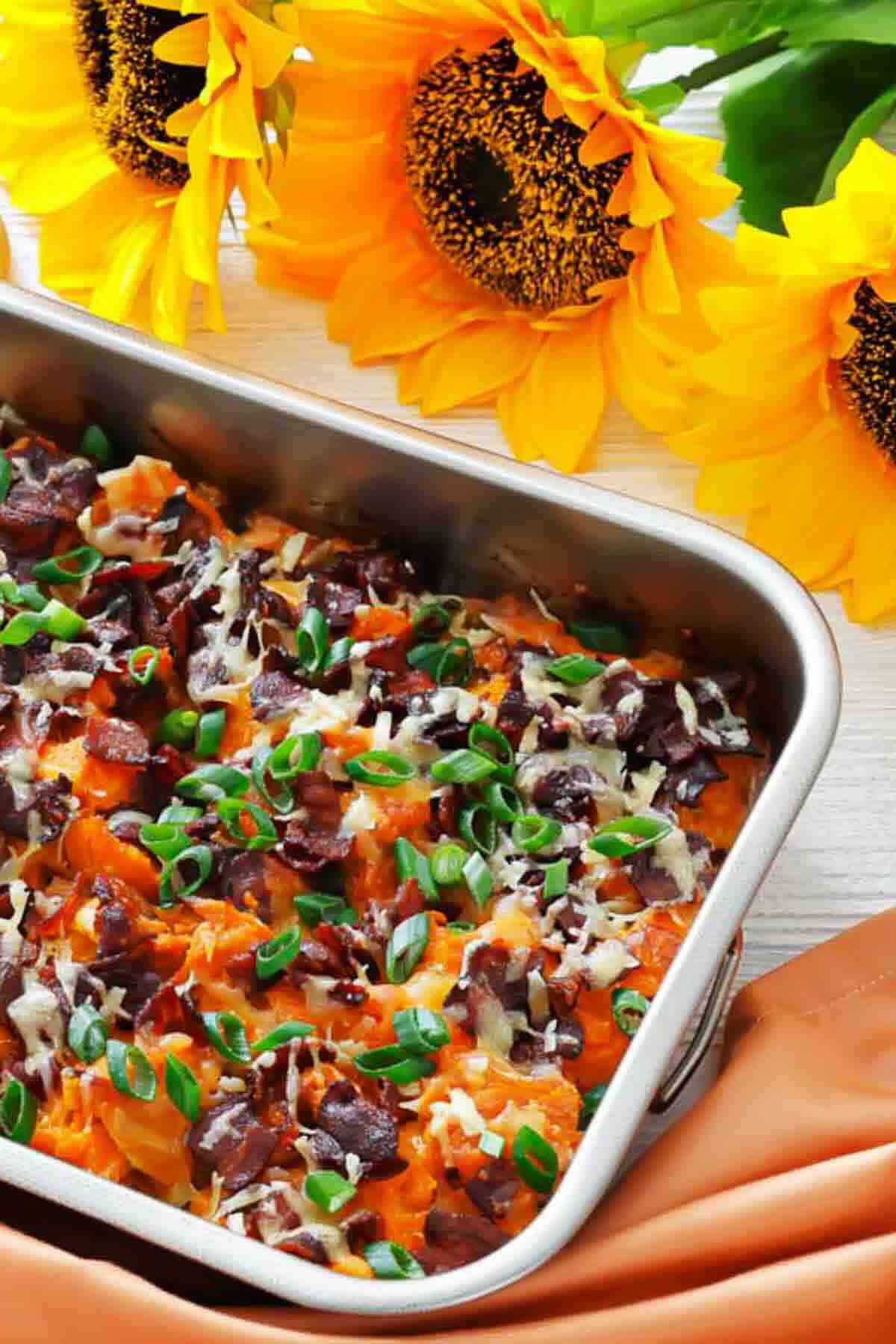 A baking dish filled with a cheesy, baked casserole topped with chopped green onions and sliced olives, set beside vibrant yellow sunflowers on a white surface.