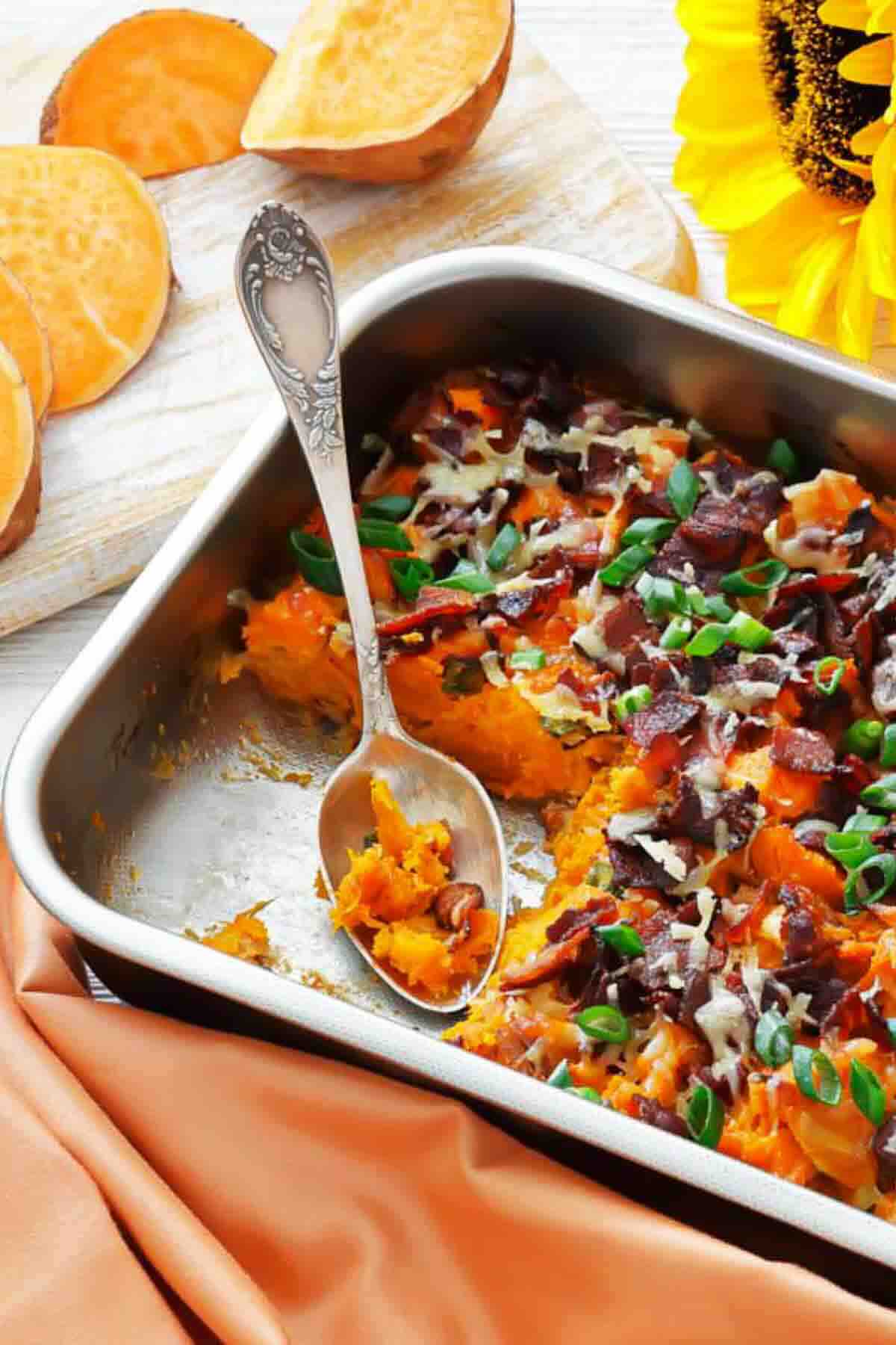 A baking dish of mashed sweet potatoes topped with bacon, cheese, and green onions, with a serving spoon and a sunflower in the background.