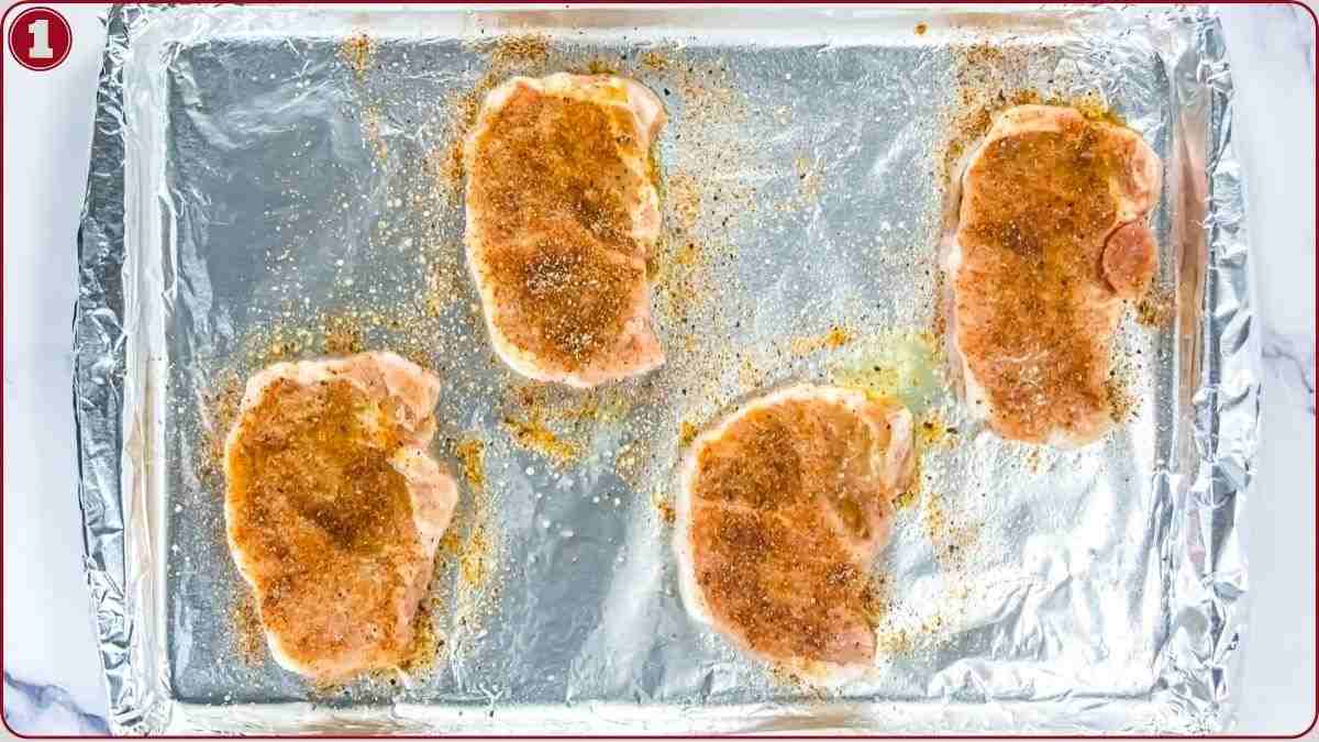Four raw, seasoned pork chops are placed on a foil-lined baking sheet, ready to be cooked.