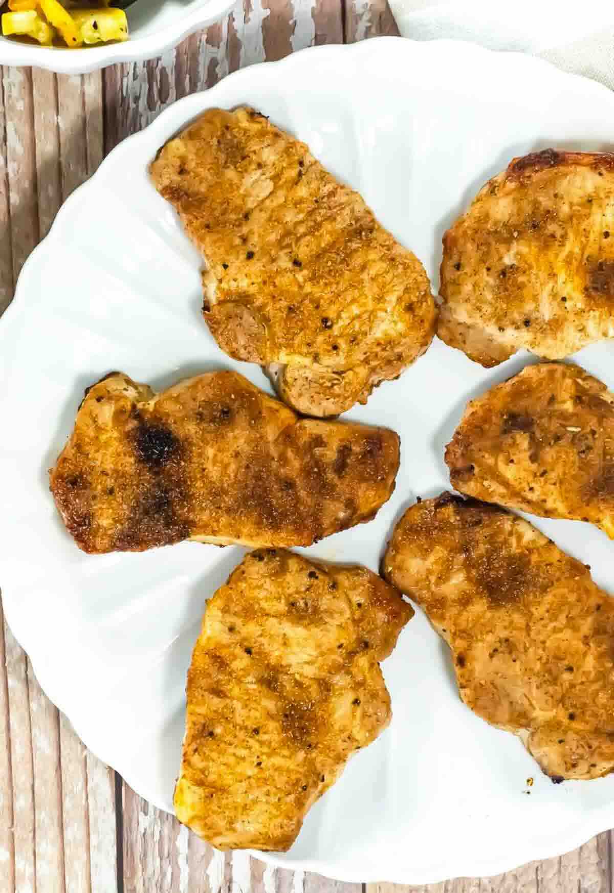 Five cooked, seasoned pork chops arranged on a white plate, viewed from above, on a wooden surface.