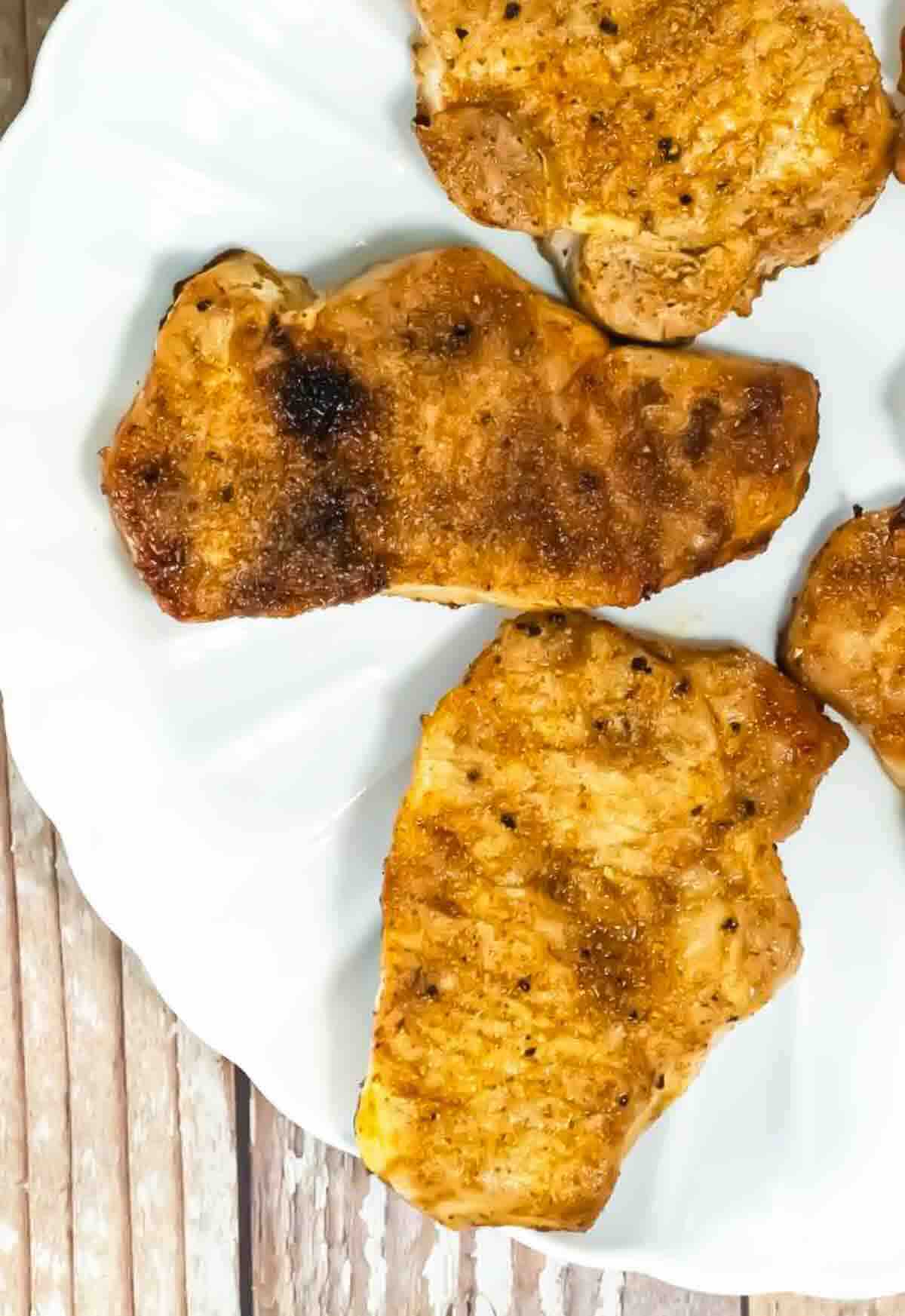 Three grilled, seasoned pork chops arranged on a white plate over a wooden surface.