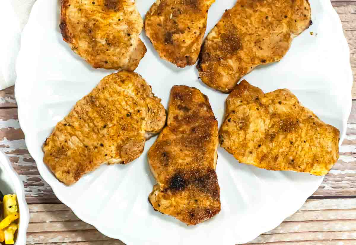 Six seasoned and cooked boneless pork chops are arranged on a white plate, displayed on a wooden surface.