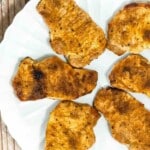 Six cooked, seasoned pork chops arranged on a white plate, viewed from above on a wooden surface.