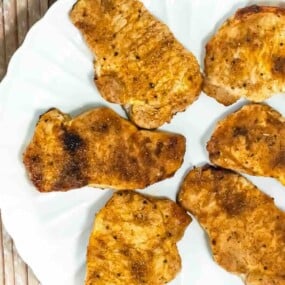 Six cooked, seasoned pork chops arranged on a white plate, viewed from above on a wooden surface.