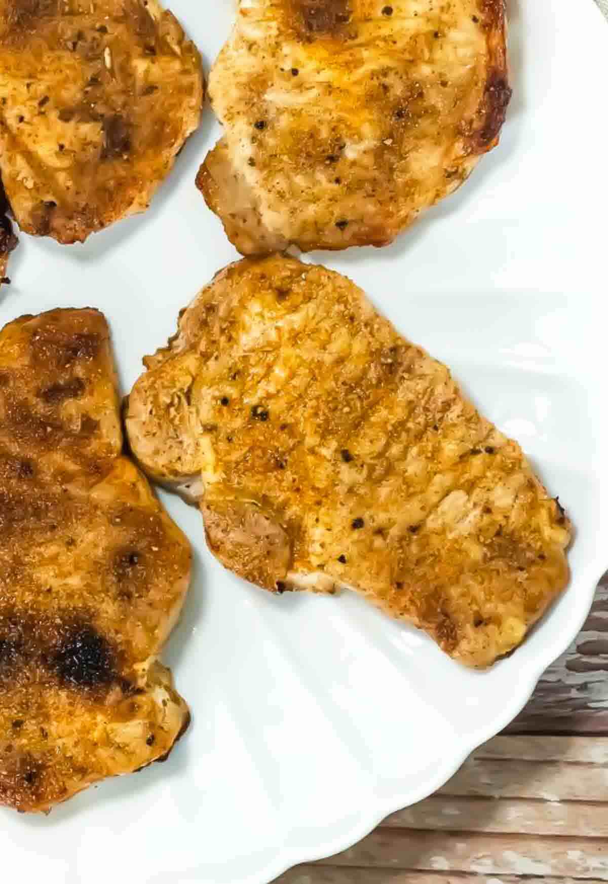 Four cooked, seasoned pork chops are arranged on a white plate, with a wooden surface partially visible underneath.