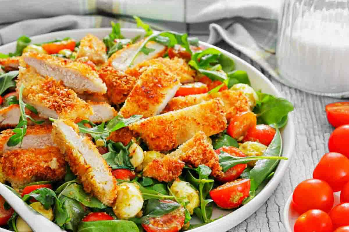 A plate of salad with cherry tomatoes, mozzarella balls, fresh greens, and sliced breaded chicken cutlets. A glass container with white liquid is in the background.