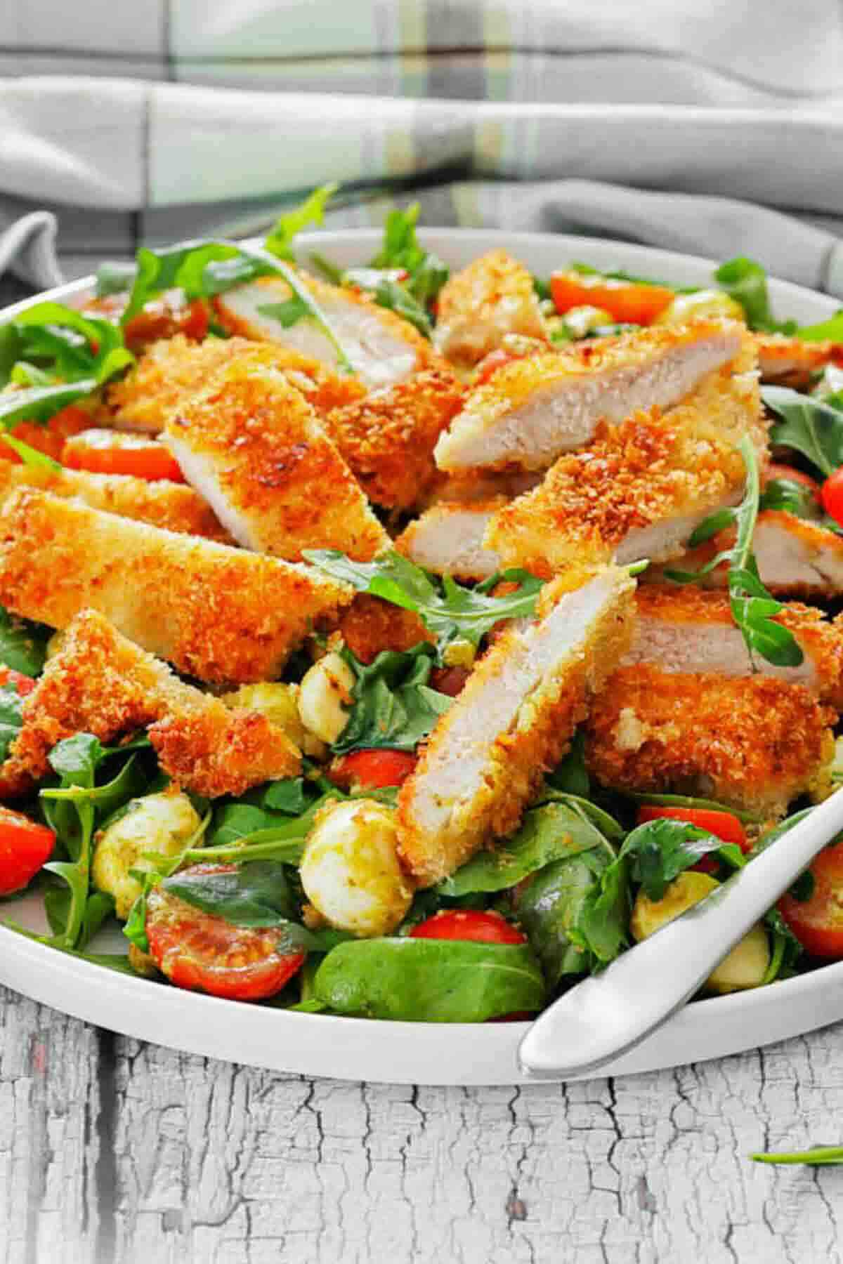A plate of breaded chicken strips on a salad with arugula, cherry tomatoes, mozzarella balls, and spinach.