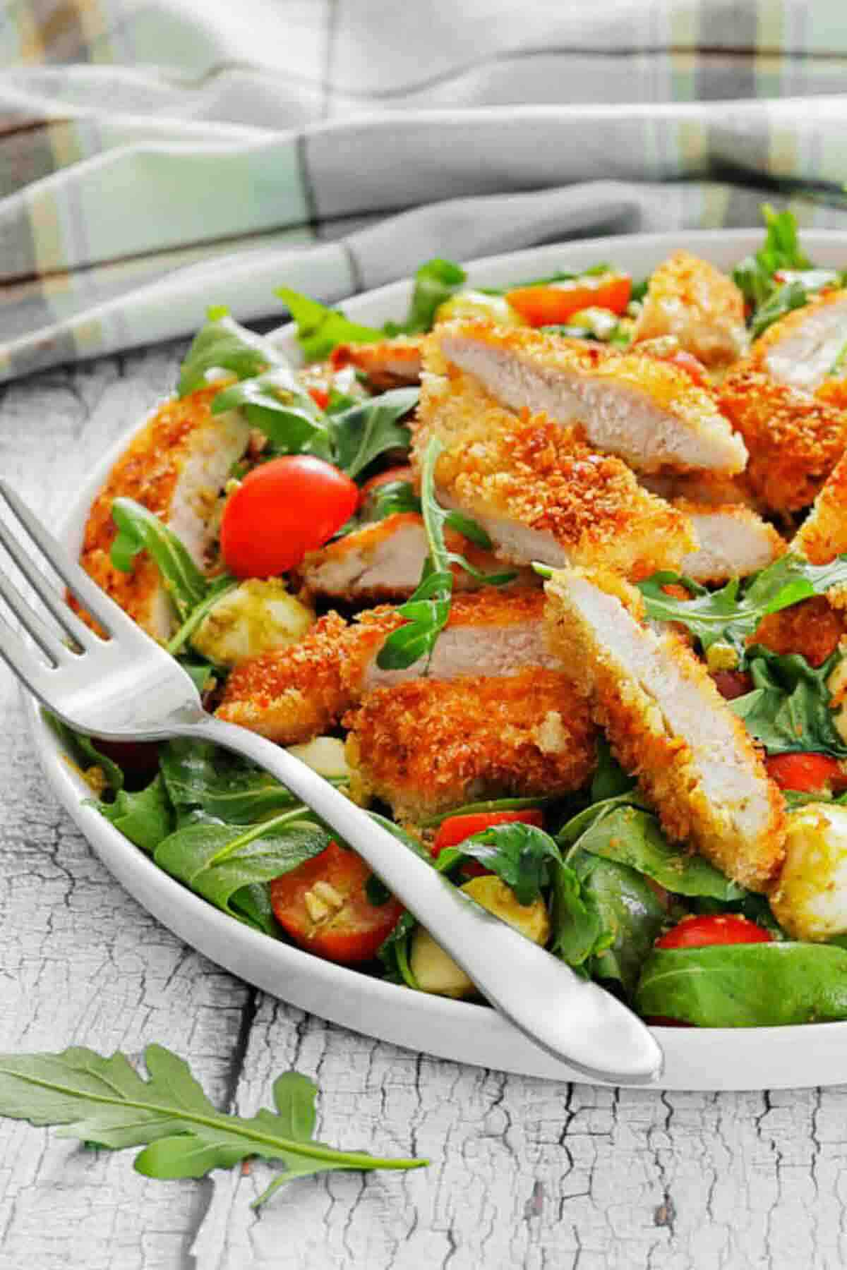 A plate of breaded chicken strips on a bed of mixed greens, cherry tomatoes, and avocado, with a fork and knife beside the food.