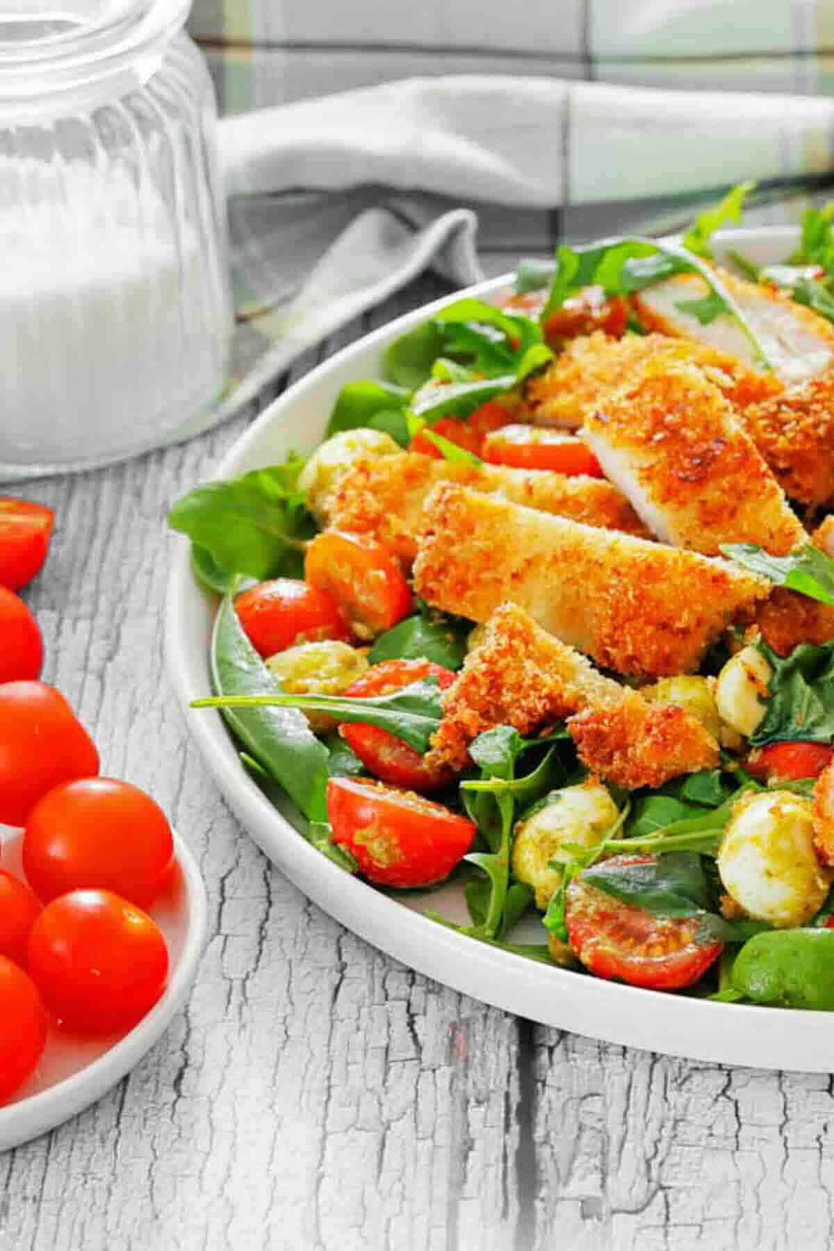 A plate of salad with breaded chicken strips, cherry tomatoes, mozzarella balls, and greens sits on a rustic wooden table beside a small bowl of cherry tomatoes.