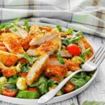 Plate of sliced breaded chicken on a green salad with cherry tomatoes, mozzarella balls, and leafy greens. A fork rests on the edge of the plate.