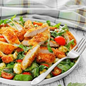 Plate of sliced breaded chicken on a green salad with cherry tomatoes, mozzarella balls, and leafy greens. A fork rests on the edge of the plate.