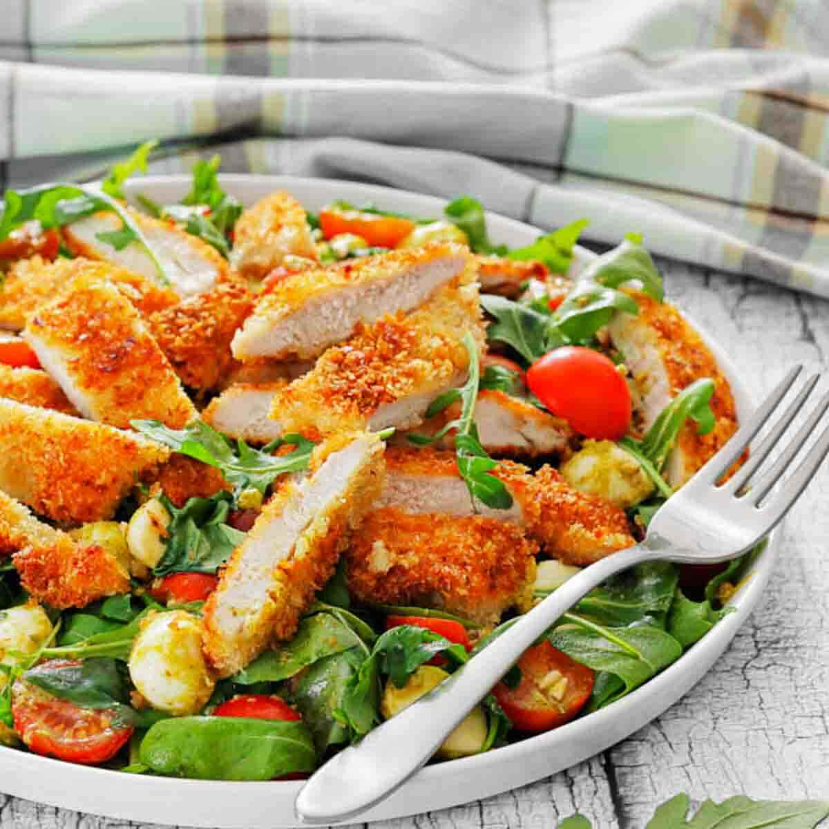 Plate of sliced breaded chicken on a green salad with cherry tomatoes, mozzarella balls, and leafy greens. A fork rests on the edge of the plate.