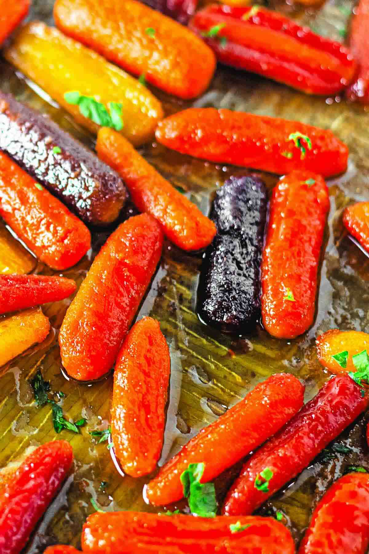 Glazed roasted baby carrots in orange, purple, and yellow colors on a baking sheet, garnished with small parsley pieces.