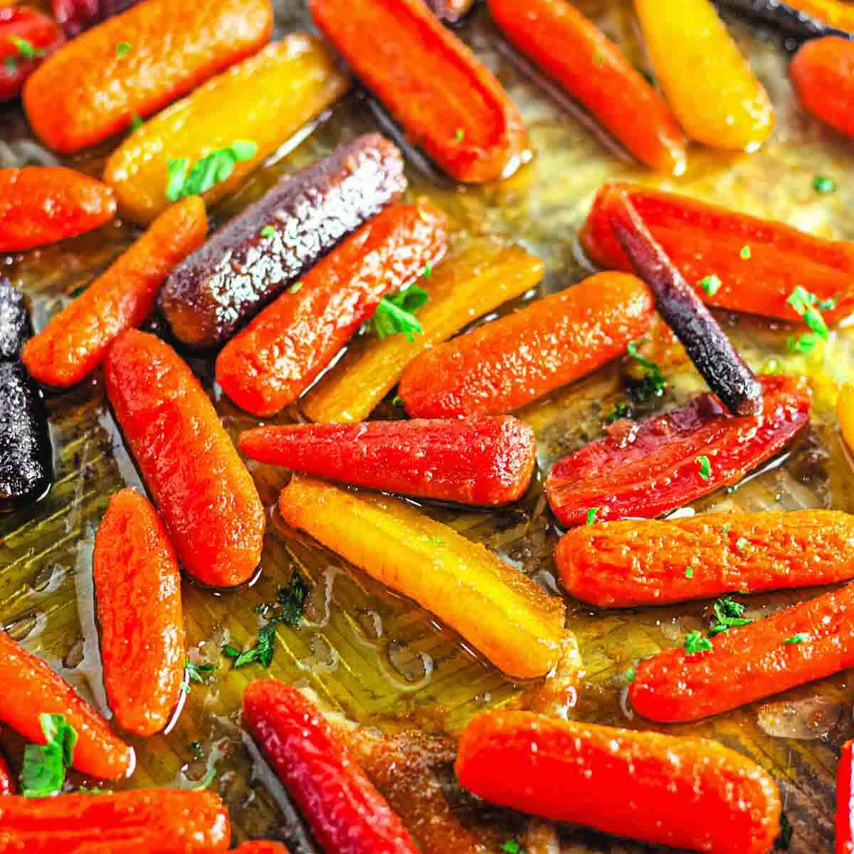 Roasted multicolored baby carrots coated in oil and garnished with chopped herbs on a baking sheet.