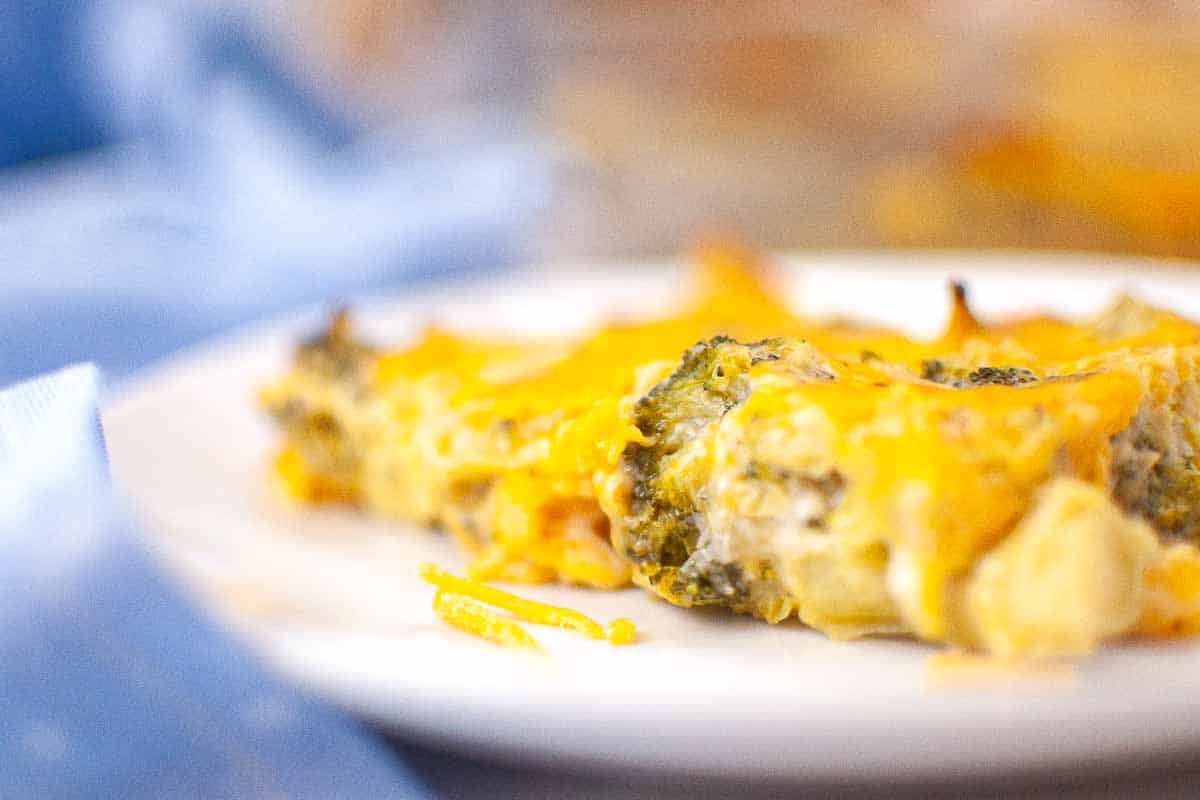 A close-up of a cheesy broccoli and potato casserole slice on a white plate, with melted cheese visible on top.