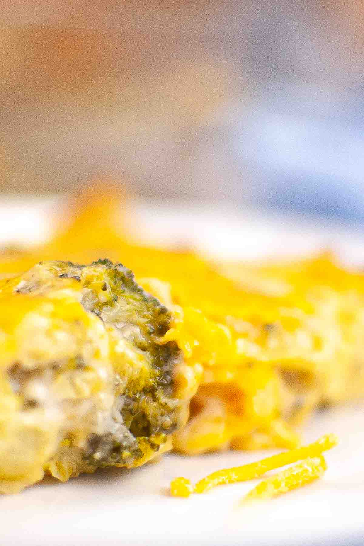Close-up of a cheesy broccoli casserole on a white plate, with melted cheese covering the top and some cheese pieces on the plate.