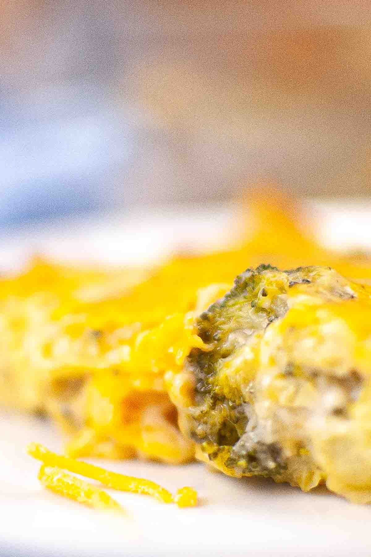Close-up of a cheesy broccoli casserole slice on a white plate, with melted cheese and broccoli pieces visible.