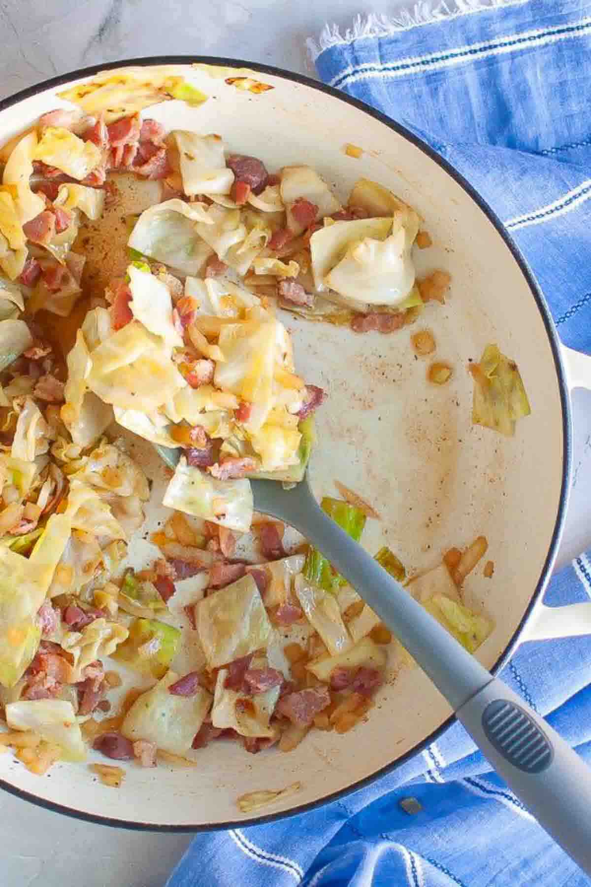 Cooked cabbage and bacon pieces in a white pot with a gray spatula, next to a blue and white striped cloth.