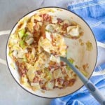 Cooked cabbage and bacon pieces in a large white pot with a spoon, sitting on a gray surface next to a folded blue cloth napkin.