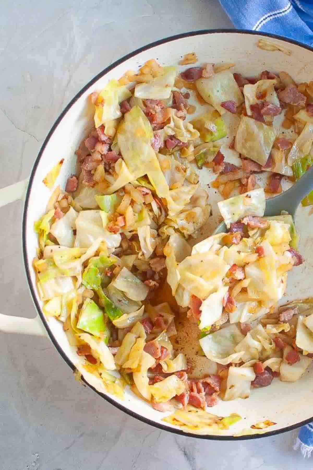 A skillet filled with sautéed cabbage and diced bacon on a light countertop with a spoon in the pan.