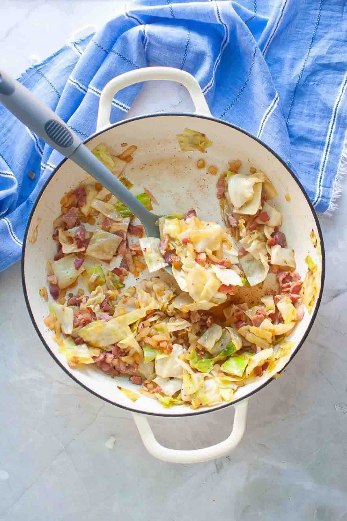White pot with sautéed cabbage, bacon, and onions on a light surface, next to a blue kitchen towel. A spoon rests in the pot.