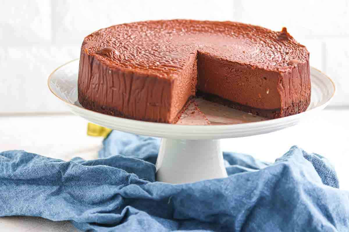 Espresso cheesecake with a slice removed, displayed on a white cake stand with a blue cloth underneath.