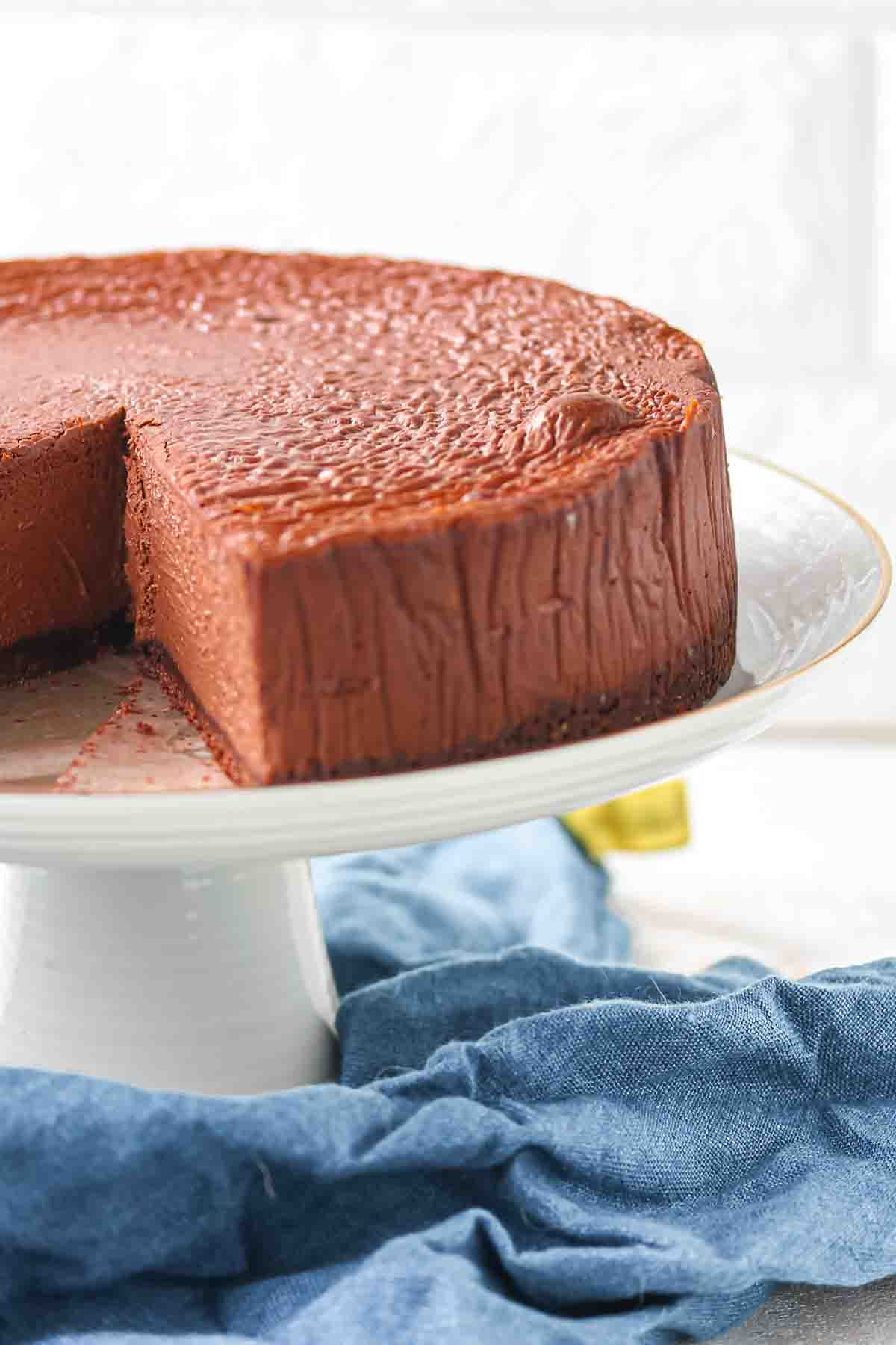 A chocolate cheesecake with a slice removed sits on a white cake stand, which is placed on a blue cloth.