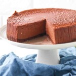 A chocolate cheesecake with a slice removed sits on a white cake stand, revealing its smooth and creamy texture.