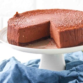 A chocolate cheesecake with a slice removed sits on a white cake stand, revealing its smooth and creamy texture.