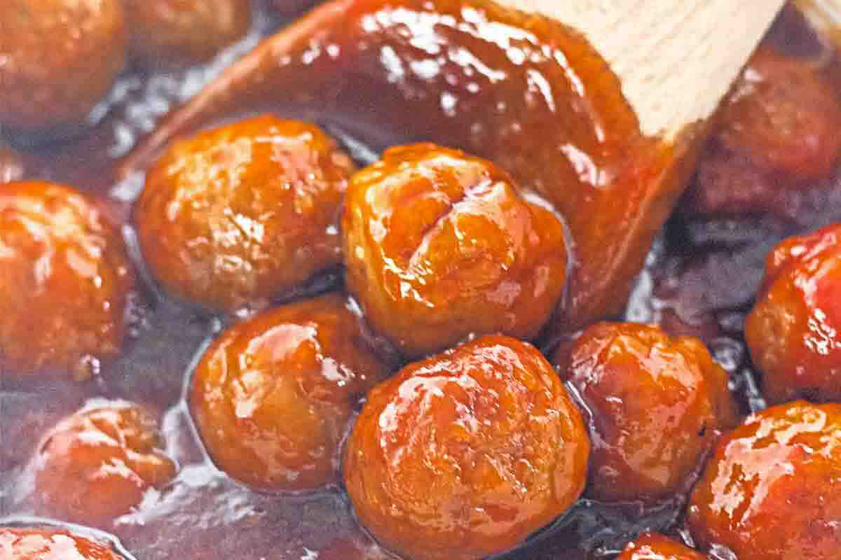 Meatballs coated in a glossy red sauce are being stirred with a wooden spoon.