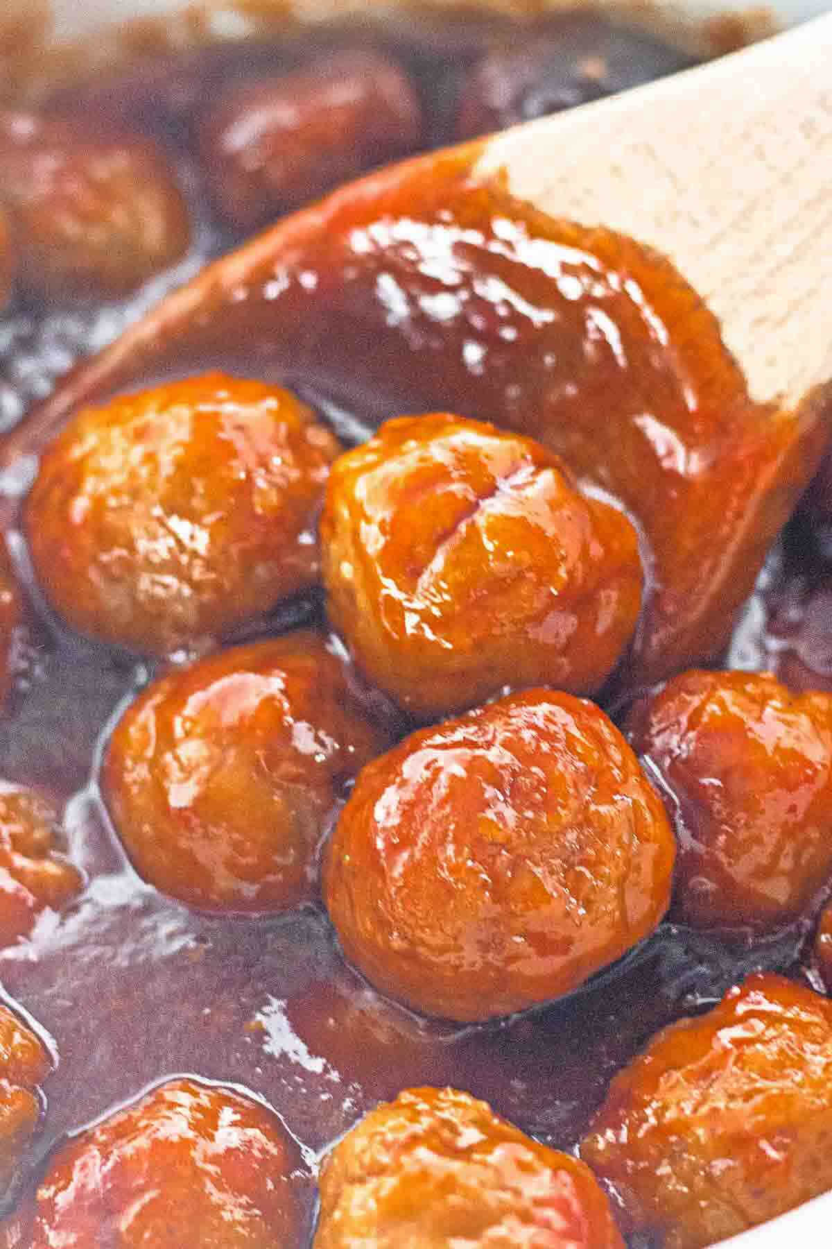 Meatballs coated in a thick, shiny brown sauce are being stirred with a wooden spoon.