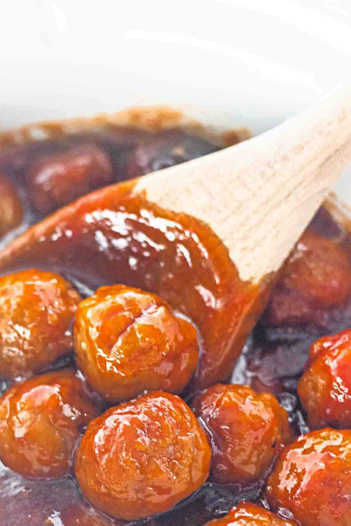 Wooden spoon stirring glazed meatballs in a thick, reddish-brown sauce.
