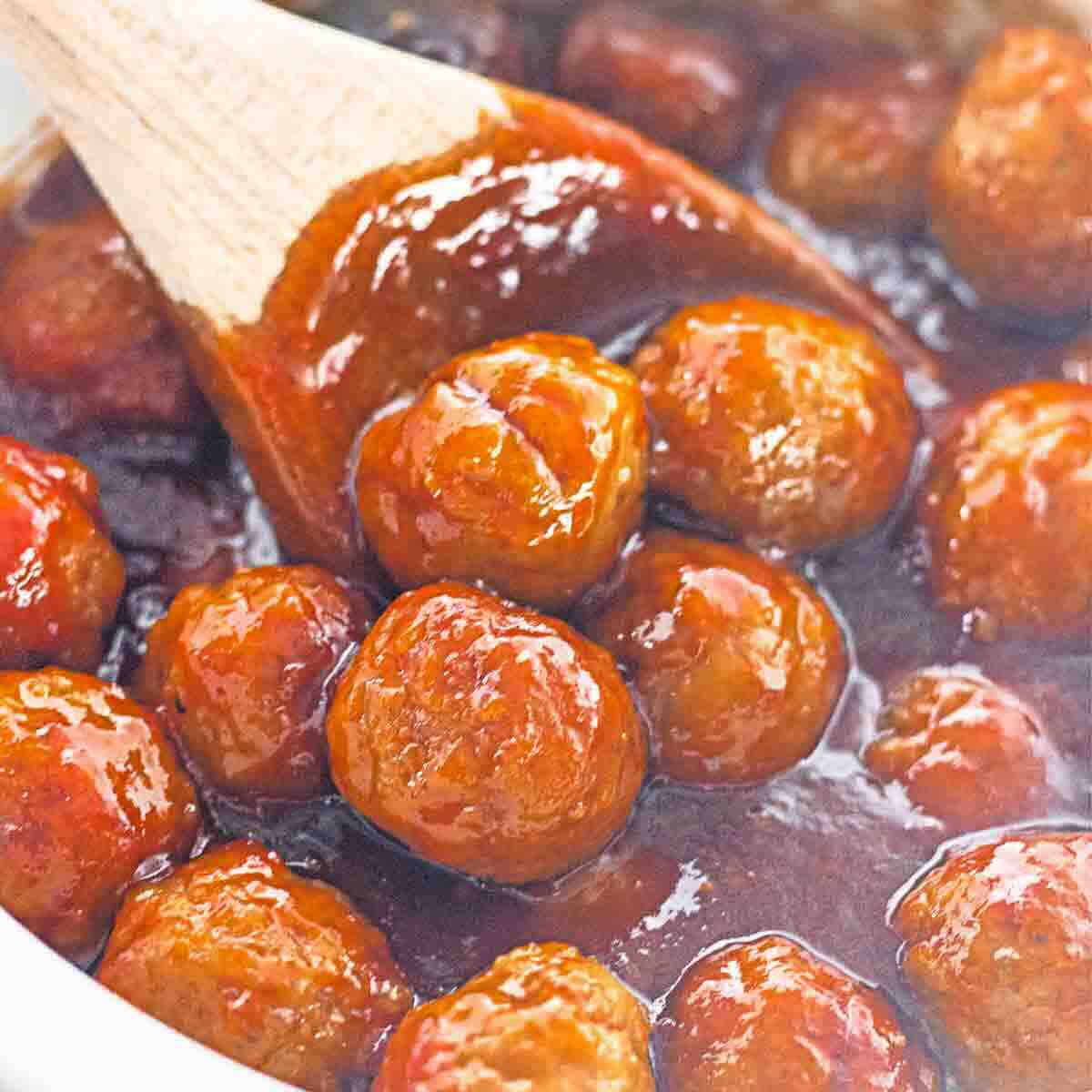 Meatballs coated in a thick, glossy brown sauce are being stirred with a wooden spoon in a pot.