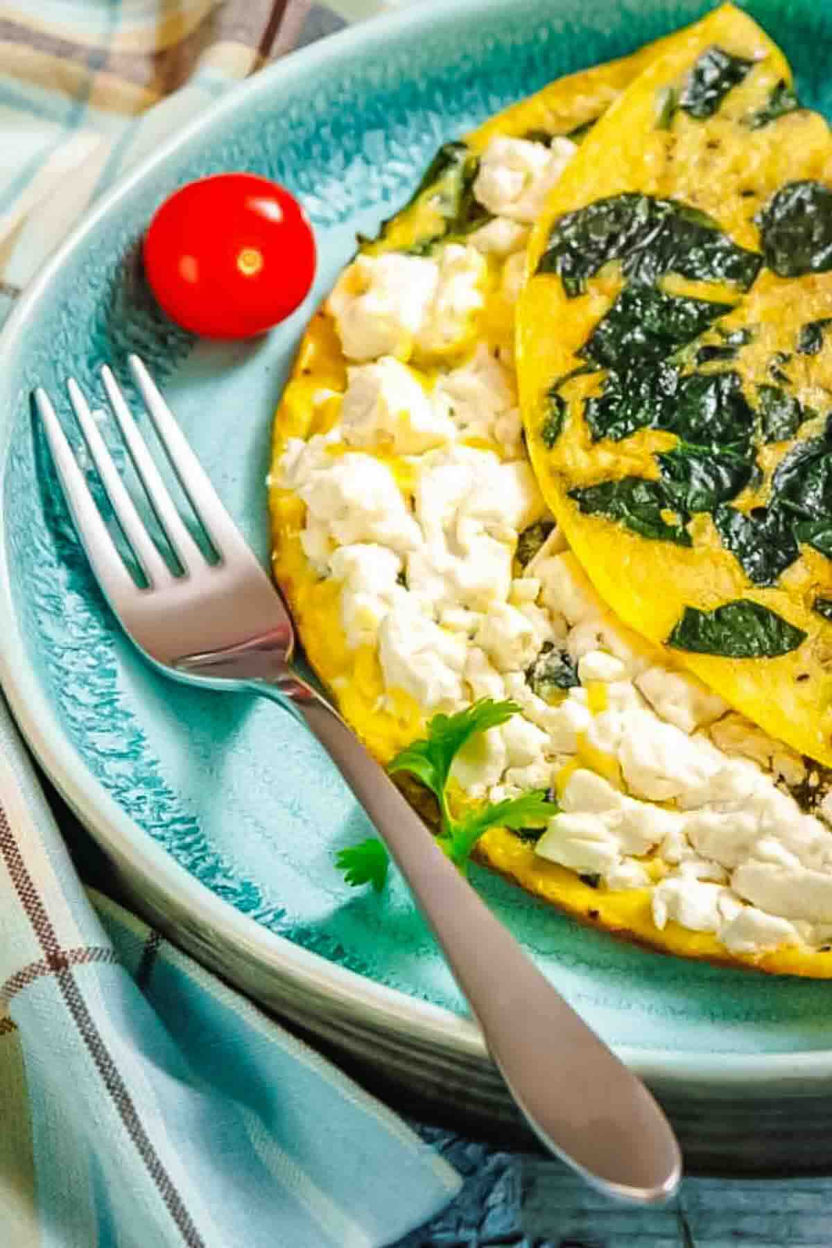 A folded omelet filled with spinach and white cheese on a blue plate, served with a cherry tomato and a fork.