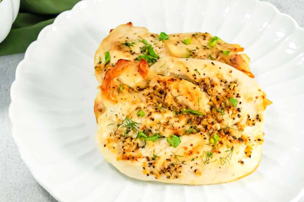 Two cooked chicken breasts seasoned with herbs and pepper on a white scalloped plate.