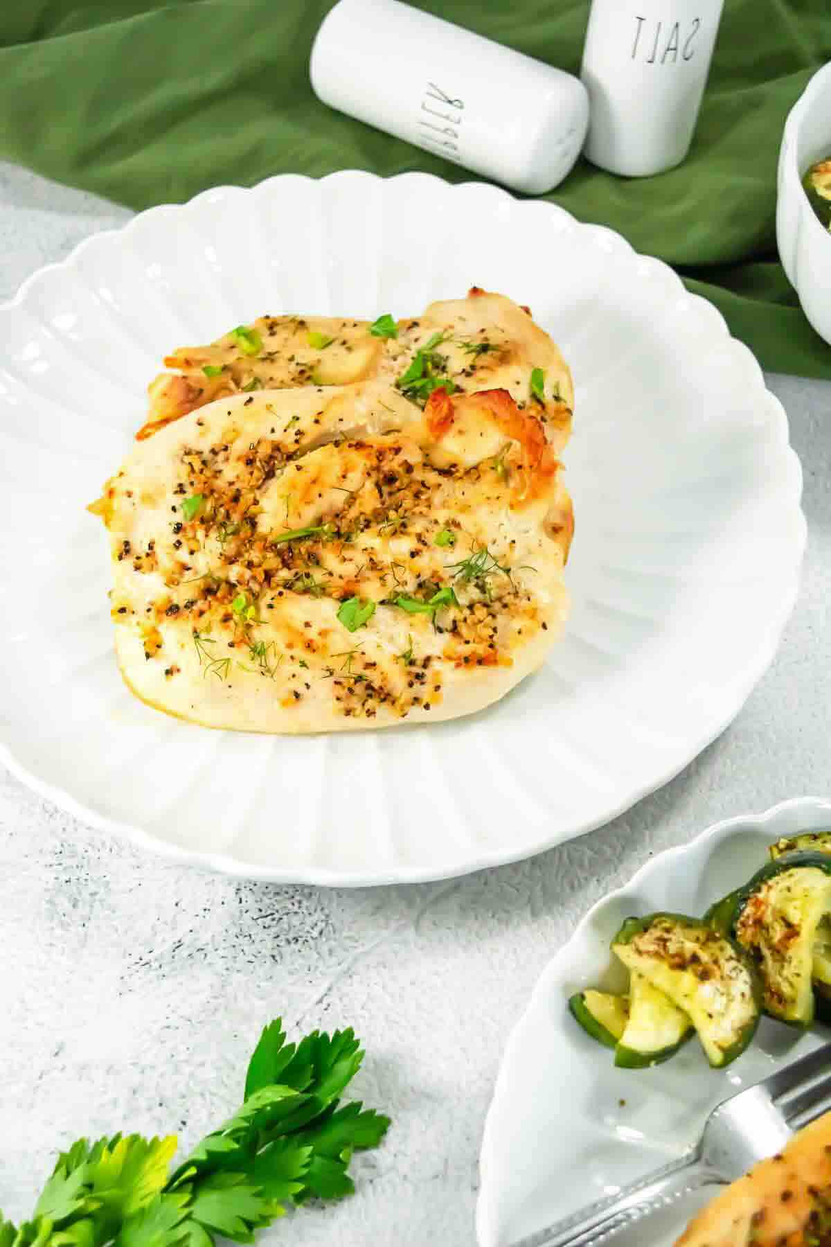 Two cooked chicken breasts topped with herbs and spices on a white plate, served with a side of sliced zucchini.