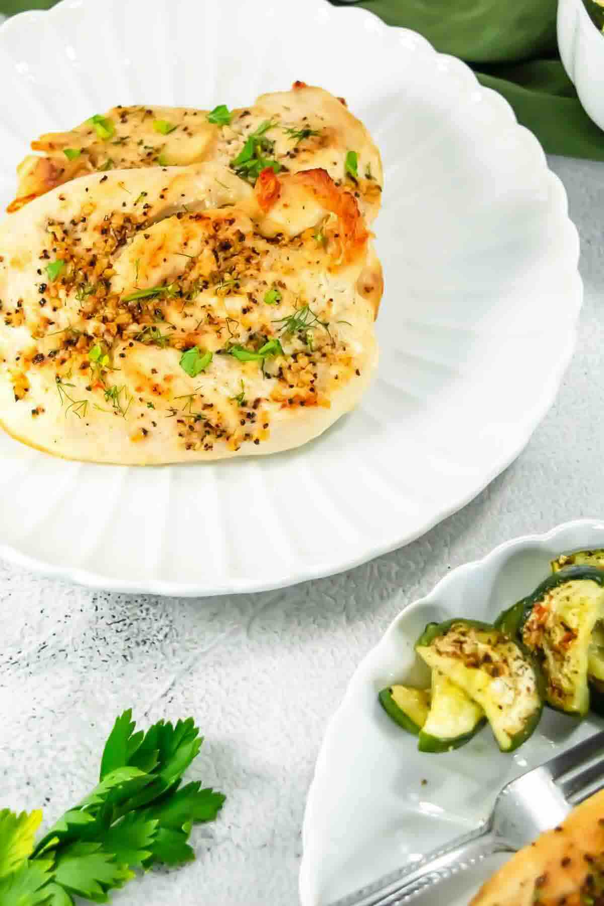Two seasoned chicken breasts garnished with herbs on a white plate, served with a side of sliced zucchini.